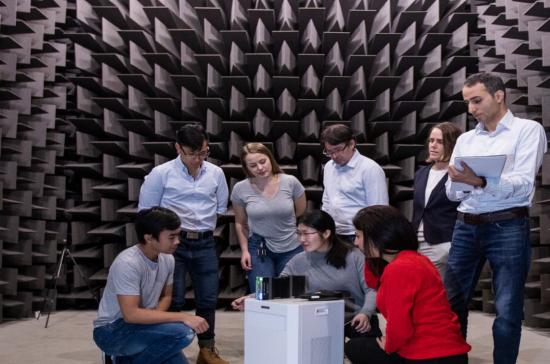 A group of seven academic researchers examining a device in a sound-proofed laboratory.