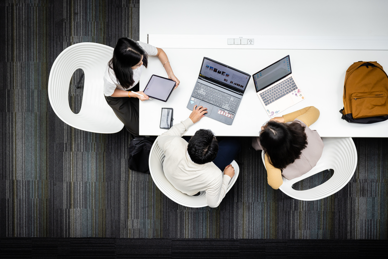 Three students sit around a table on laptops and tablet