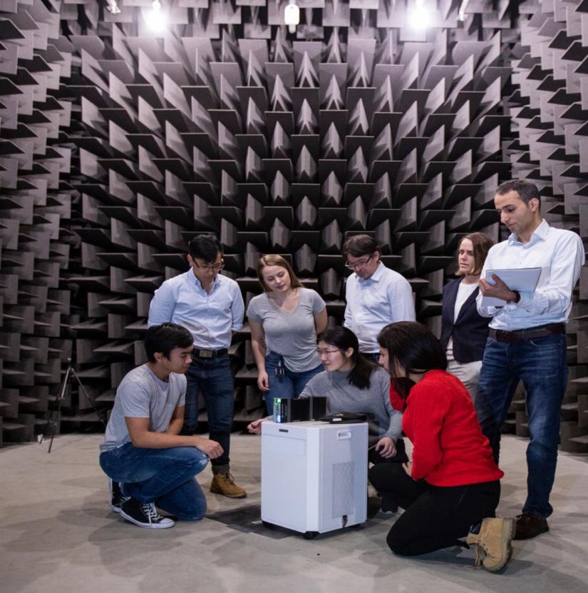 A group of seven academic researchers examining a device in a sound-proofed laboratory.